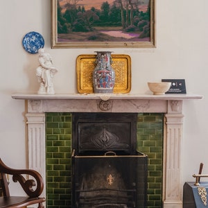May include: A white marble fireplace with a green tiled surround and a black metal fire grate. A white porcelain statue, a gold framed landscape painting, and a blue and white porcelain vase are on the mantel.