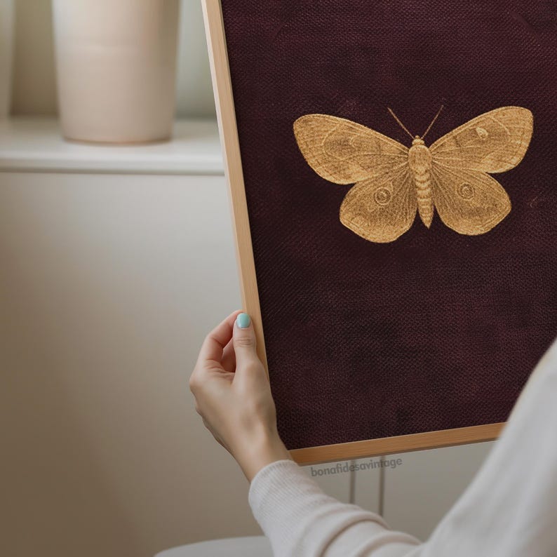 May include: A framed artwork featuring a detailed gold-colored butterfly illustration on a dark burgundy background. The butterfly is centered, with its wings spread. The frame is a light wood color. The artwork is held by a person.