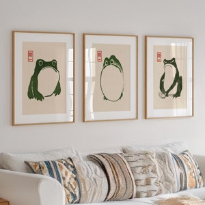 May include: Three framed prints with green frog illustrations. The frogs are in different poses, one is sitting, one is standing, and one is in a circle. The frames are light wood and have a white mat.