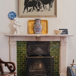 May include: A white marble fireplace with a green tiled surround and a black metal fireplace screen. A vase with a colorful floral design sits on the mantel above the fireplace, along with a framed black and white print of horses.