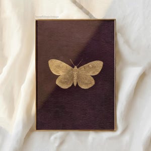 May include: A framed print featuring a gold-colored butterfly illustration on a dark purple background. The butterfly is centered, with detailed wing patterns. The frame is a thin, gold metal. The print is displayed on a white fabric surface.