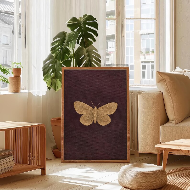 May include: A framed art print featuring a gold-colored butterfly against a deep burgundy background. The artwork is displayed in a room with natural light, a large plant, and wooden furniture, creating a warm and inviting atmosphere.