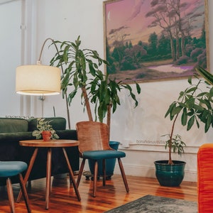 May include: A living room interior with a green sofa, two blue chairs, a wooden table, a floor lamp, and two potted plants. A framed landscape painting hangs on the wall above the sofa. A rug covers the wooden floor.