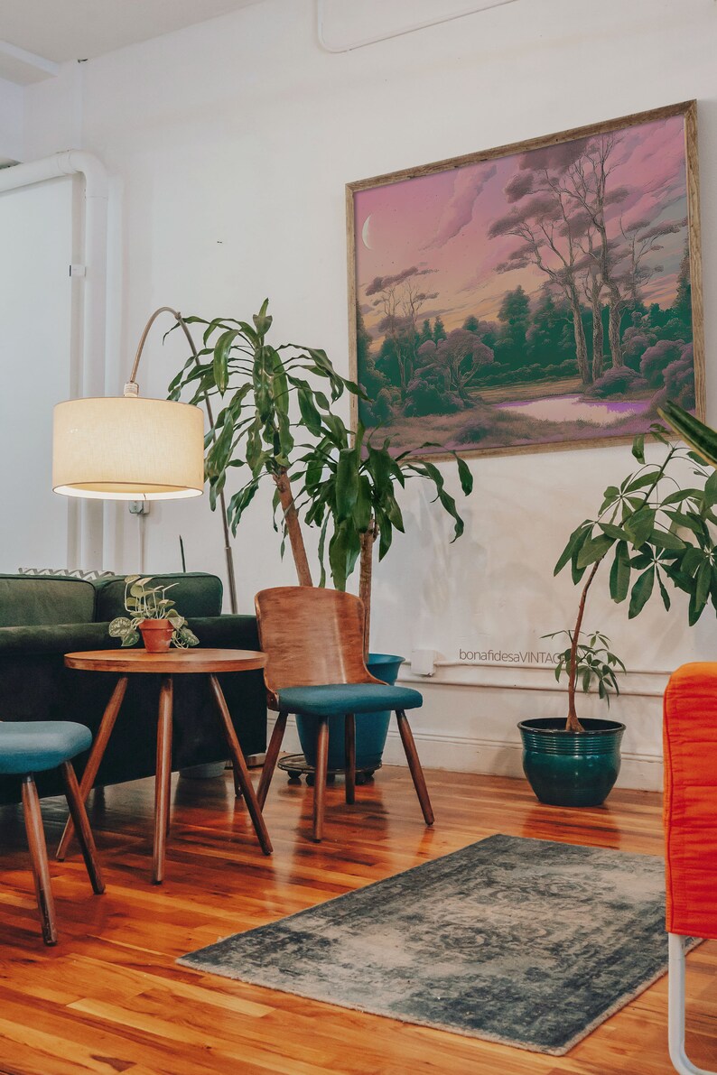 May include: A living room interior with a green sofa, two blue chairs, a wooden table, a floor lamp, and two potted plants. A framed landscape painting hangs on the wall above the sofa. A rug covers the wooden floor.