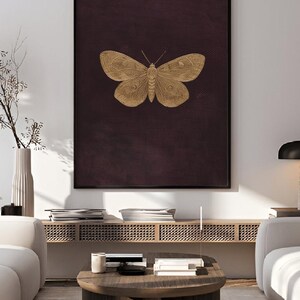 May include: A framed wall art print featuring a gold-colored butterfly illustration on a dark burgundy background. The artwork is displayed in a modern living room setting, with the text "bonafidesavintage" in the top left corner.