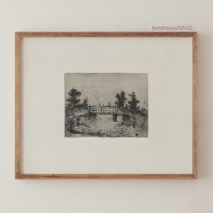 May include: A framed black and white etching of a small bridge over a stream with trees on either side. The etching is in a light wood frame.
