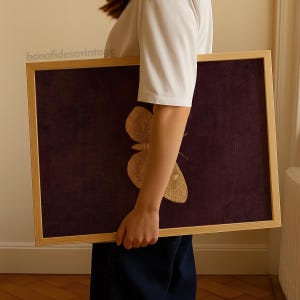 May include: A framed artwork featuring a detailed illustration of a butterfly. The butterfly is in shades of gold against a dark purple textured background. The frame is made of light-colored wood. The text "bonafidesavintage" is in the upper left corner.