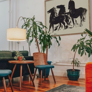 May include: A black and white abstract art print of three horses running, framed in a light wood frame, hanging on a white wall above a green sofa and a wooden coffee table with a potted plant on top.