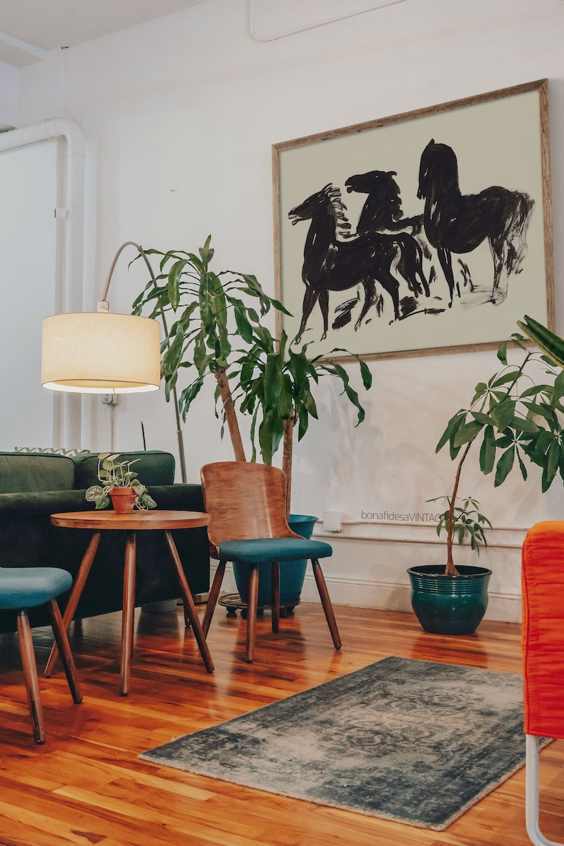 May include: A black and white abstract art print of three horses running, framed in a light wood frame, hanging on a white wall above a green sofa and a wooden coffee table with a potted plant on top.