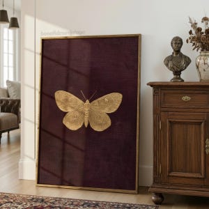 May include: A framed artwork featuring a gold-colored butterfly on a deep burgundy textured background. The butterfly is centrally positioned, with detailed wing patterns. The frame is a thin, gold color. The artwork is leaning against a white wall.