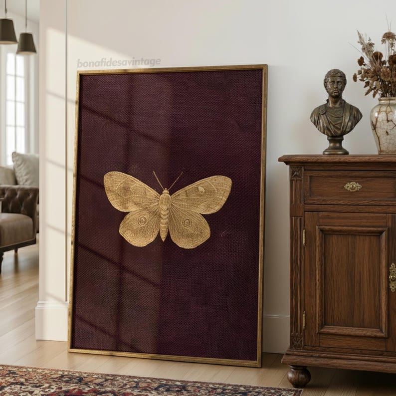 May include: A framed artwork featuring a gold-colored butterfly on a deep burgundy textured background. The butterfly is centrally positioned, with detailed wing patterns. The frame is a thin, gold color. The artwork is leaning against a white wall.