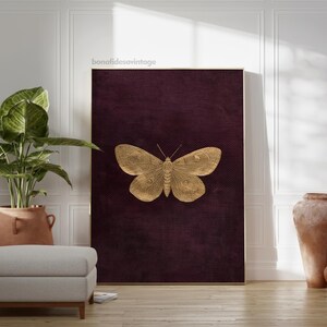 May include: A framed print featuring a gold butterfly on a dark burgundy textured background. The butterfly is detailed with intricate wing patterns. The print is displayed in a room with a plant and a neutral-toned ottoman.