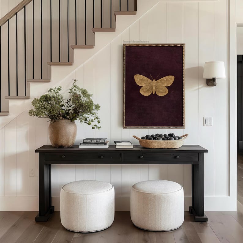May include: A dark wood console table with two drawers, topped with books, a woven basket, and a large vase of greenery. Two upholstered ottomans sit in front of the table. A framed butterfly art piece hangs on the white paneled wall.