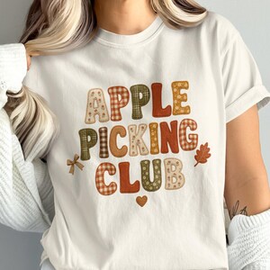 Op de afbeelding: Een crèmekleurig T-shirt met korte mouwen en de tekst APPLE PICKING CLUB in een veelzijdig lettertype met aardetinten. Het ontwerp bevat kleine decoratieve elementen zoals een bruine strik, een klein hartje en een oranje herfstblad.