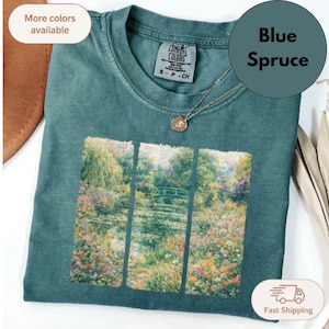 Puede incluir: Una camiseta de color azul abeto con un estampado gr&aacute;fico de un paisaje de jard&iacute;n con un puente dividido en tres paneles verticales. La camiseta tiene cuello redondo y una etiqueta que dice Comfort Colors con la talla S P CH visible.