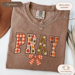 Op de afbeelding: Een espresso-kleurig t-shirt met het woord PRAY in grote blokletters. Elke letter heeft een uniek patroon: rood gingham, herfstbladeren, bloemenprint en bruine ruit. Een rode gingham strik is onder het woord PRAY geprint.