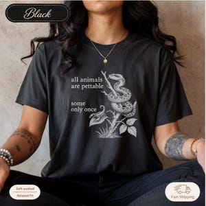Peut inclure: Un t-shirt noir à manches courtes avec une illustration graphique blanche représentant un serpent enroulé autour d'une branche et un scorpion en dessous. Le texte indique "all animals are pettable some only once". Col rond et coupe décontractée.