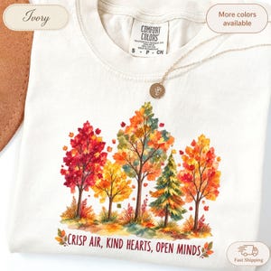 May include: An ivory t-shirt featuring a watercolor graphic of autumn trees in shades of red, orange, yellow, and green. Below the trees, the text reads "CRISP AIR, KIND HEARTS, OPEN MINDS" in a dark font. A gold-toned necklace rests on the neckline.