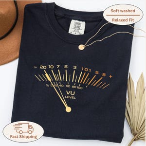 Puede incluir: Una camiseta azul marino con un diseño de medidor VU dorado. La camiseta tiene un lavado suave y un ajuste relajado. Un collar dorado con un pequeño colgante está sobre la camiseta. Un sombrero marrón y hojas de palma secas están en el fondo.
