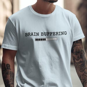 May include: A light blue t-shirt with the text "BRAIN BUFFERING" above a loading bar graphic. The shirt is a crew neck style and appears to be made of a soft material.