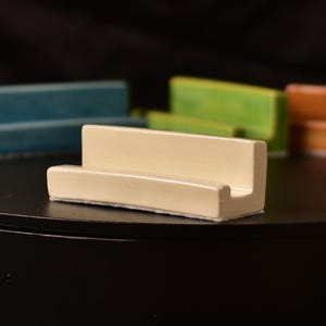 Puede incluir: Un soporte para teléfono de cerámica color crema con una base rectangular y una parte trasera elevada. El soporte está sobre una superficie negra, con otras piezas de cerámica coloridas en el fondo.