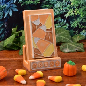 Puede incluir: Un soporte para teléfono de cerámica con temática de Halloween en color naranja claro. El soporte presenta un diseño de caramelos de maíz y la palabra "TREAT" en la base. Caramelos de maíz y una pequeña calabaza están esparcidos.