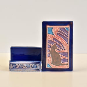 May include: Two ceramic pieces: a dark blue rectangular holder and a dark blue rectangular tile with a cat silhouette. The tile features a cat silhouette in brown, set against a backdrop of pink, purple, and blue abstract shapes.