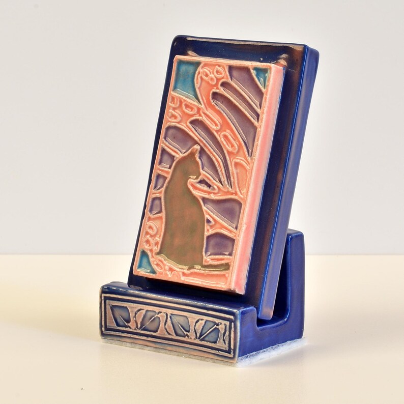 May include: A blue ceramic phone stand with a rectangular tile featuring a cat silhouette and abstract design in shades of brown, pink, purple, and blue. The stand has a decorative base with a leaf pattern.