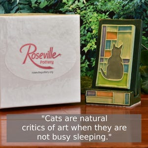 May include: A ceramic cat figurine in a stained-glass style, with a dark green frame and a gray cat silhouette. The figurine is displayed next to a white box with the "Roseville Pottery" logo and the text "Cats are natural critics of art when they are not busy sleeping."