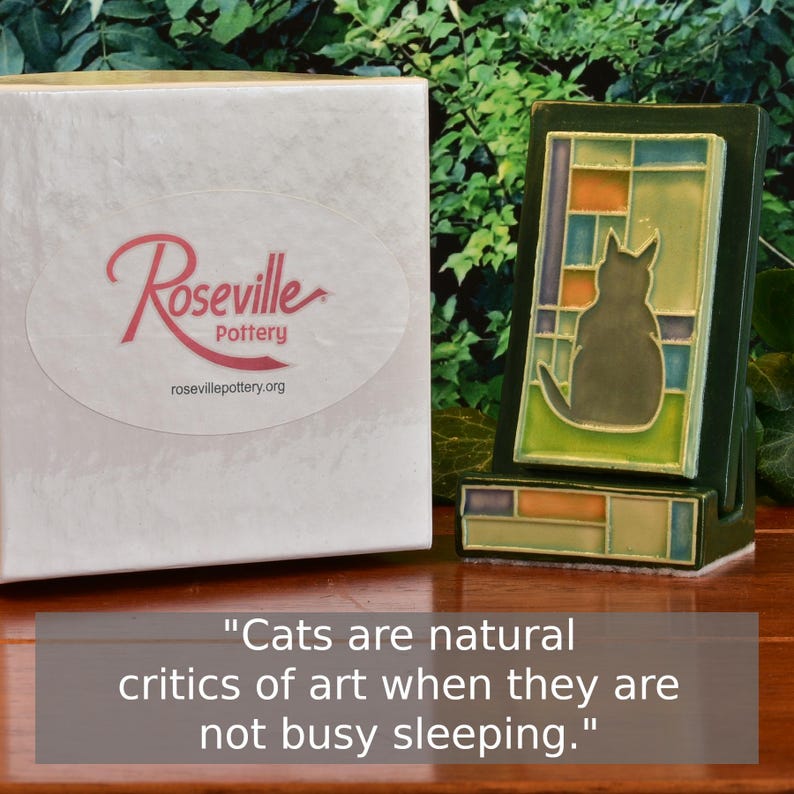 May include: A ceramic cat figurine in a stained-glass style, with a dark green frame and a gray cat silhouette. The figurine is displayed next to a white box with the "Roseville Pottery" logo and the text "Cats are natural critics of art when they are not busy sleeping."