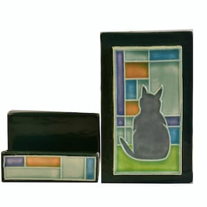 May include: Two-piece ceramic set featuring a dark green rectangular box and a rectangular tile with a cat silhouette. The tile has a stained-glass window design with blue, orange, and green squares. The cat is gray.