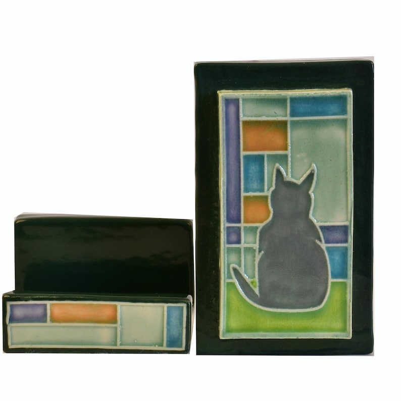 May include: Two-piece ceramic set featuring a dark green rectangular box and a rectangular tile with a cat silhouette. The tile has a stained-glass window design with blue, orange, and green squares. The cat is gray.