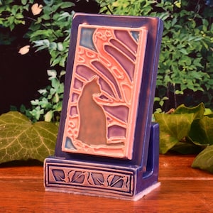 May include: A blue ceramic cat-themed tile holder. The tile features a cat silhouette in shades of brown and purple, set against a backdrop of pink, orange, and blue. The base has a decorative leaf design.