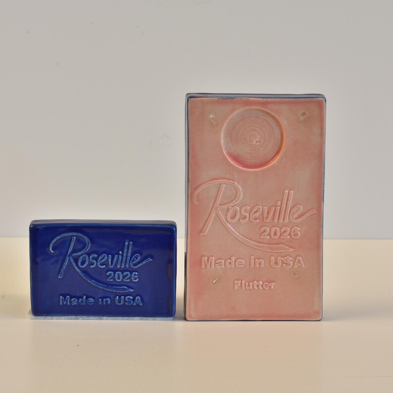 May include: Two Roseville pottery pieces. A small, rectangular, cobalt blue piece and a larger, rectangular, pink piece. Both pieces have the Roseville name and "2026" embossed on them. The pink piece also has "Made in USA" and "Flutter" embossed.
