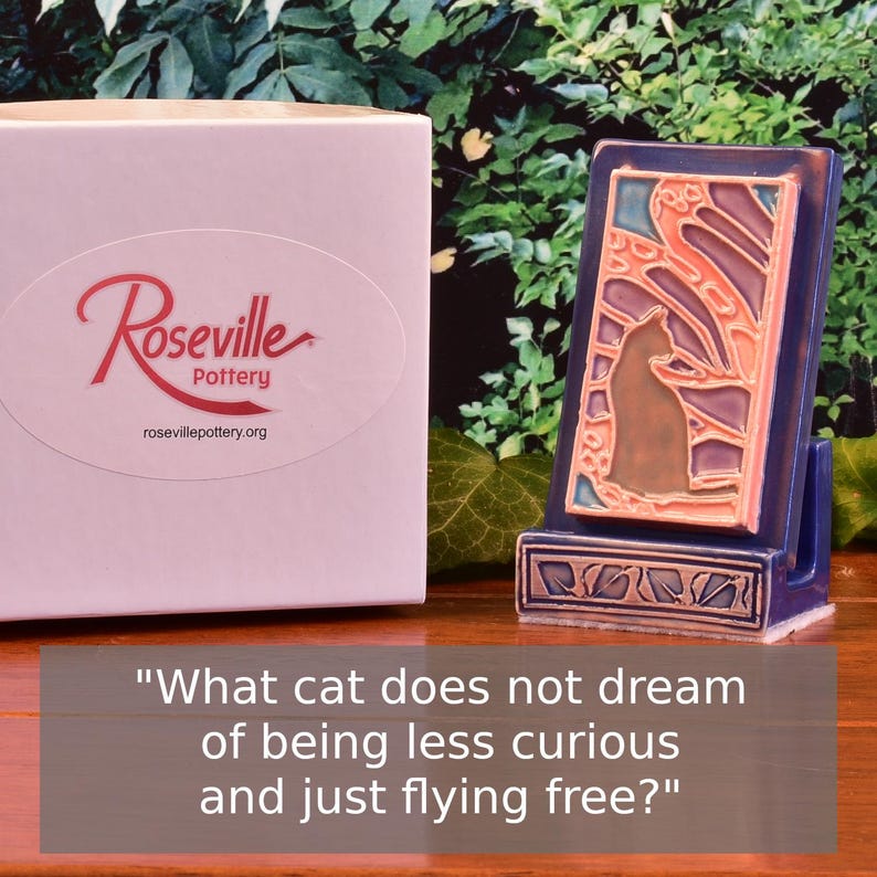 May include: A ceramic cat-themed art piece with a blue frame and a cat silhouette. The piece is displayed on a wooden surface next to a box with the "Roseville Pottery" logo and the text "What cat does not dream of being less curious and just flying free?"