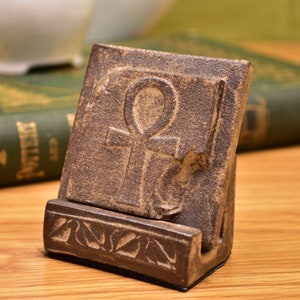 Op de afbeelding: Een bruine keramische telefoonstandaard met een Ankh-symbool dat in de voorkant is gegraveerd. De standaard heeft een decoratief gegraveerd ontwerp rond de basis.