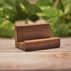 May include: A small, rectangular brown wooden stand featuring a horizontal slot designed to hold an object upright. The stand has a smooth, natural wood grain finish and a simple L-shaped profile with a flat base and a vertical back support.