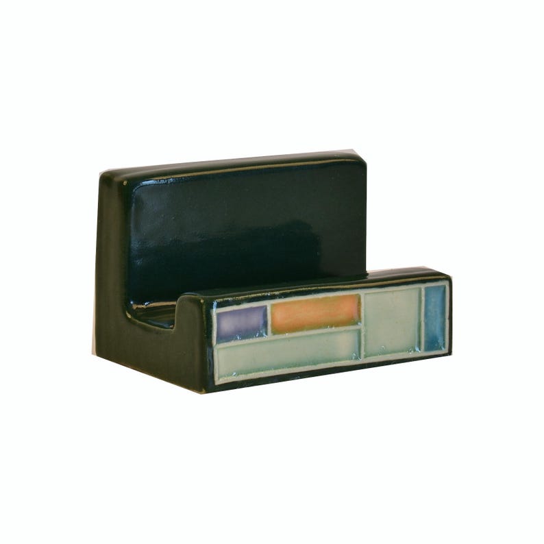 May include: A dark green ceramic wall hook with a rectangular back and a horizontal front section. The front section features a mosaic design with rectangular tiles in shades of blue, orange, and green. The hook is designed for hanging items.