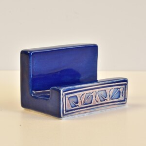 May include: A cobalt blue ceramic soap dish with a rectangular shape. The front features a decorative border with a leaf design in silver and light blue. The dish has a glossy finish and is designed to hold a bar of soap.