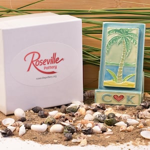 May include: A ceramic tile featuring a palm tree design, displayed on sand with seashells. The tile has a blue frame, a green palm tree, and a blue ocean. A white box with the "Roseville Pottery" logo is on the left.
