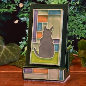 Może przedstawiać: Ciemnozielona ceramiczna ramka na zdjęcia z motywem kota. Ramka posiada witrażowy wzór z szarą sylwetką kota. Witraż ma pomarańczowe, niebieskie i fioletowe akcenty. Ramka jest prostokątna z podstawą.