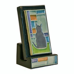 May include: A dark green ceramic phone stand with a decorative tile featuring a black cat silhouette. The tile has a stained glass design with blue, orange, and green squares. The stand is angled for viewing.