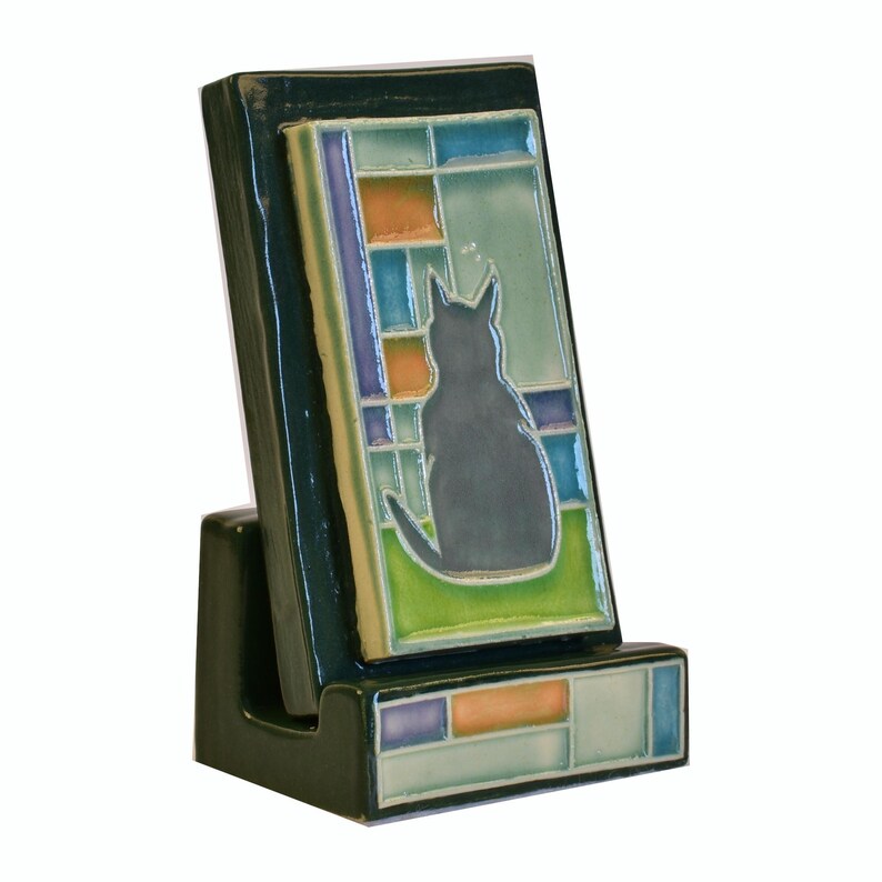 May include: A dark green ceramic phone stand with a decorative tile featuring a black cat silhouette. The tile has a stained glass design with blue, orange, and green squares. The stand is angled for viewing.