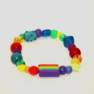 May include: A stretch bracelet featuring a variety of colourful plastic beads in shapes including spheres, stars, and a central rectangular charm with horizontal rainbow stripes in red, orange, yellow, green, blue, and purple, arranged in a circular loop.