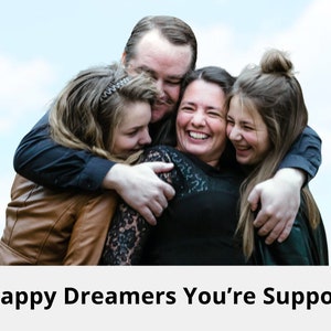 May include: A family of four, two adults and two children, are hugging each other and smiling. They are standing in front of a blue sky with white clouds.