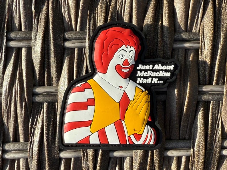 K&ouml;nnte beinhalten: Ein gestickter Aufn&auml;her mit einem Comic-Charakter mit einer roten Per&uuml;cke und einem gelb-rot gestreiften Hemd. Der Charakter h&auml;lt seine H&auml;nde zum Beten zusammen. Der Text "Just About McFuckin Had It..." ist in Wei&szlig; auf schwarzem Hintergrund geschrieben.