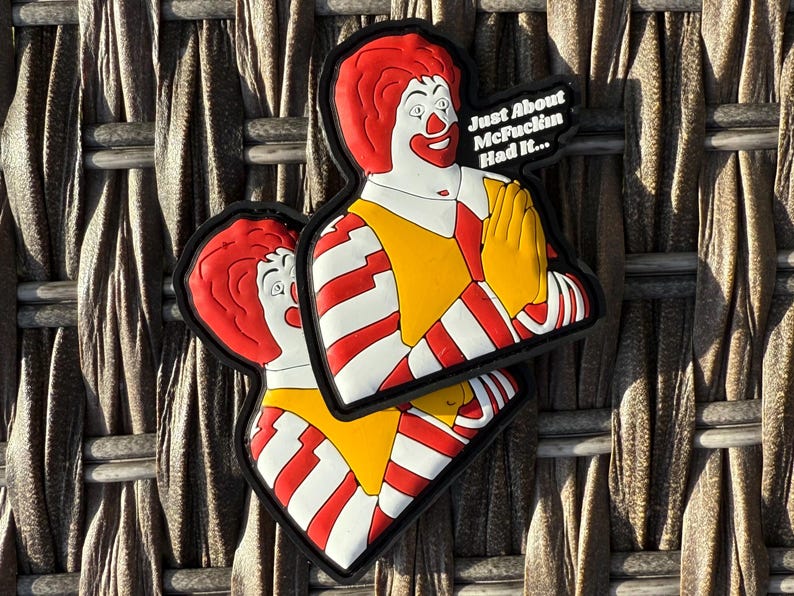 K&ouml;nnte beinhalten: Zwei rot-wei&szlig; gestreifte Patches mit einem Cartoon-Bild von Ronald McDonald. Der obere Patch hat den Text "Just About McFuckin Had It..." Die Patches befinden sich auf einem braunen gewebten Hintergrund.