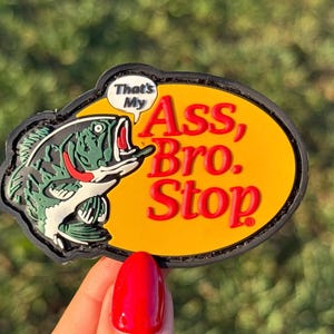 Könnte beinhalten: Ein origineller Aufnäher mit einer grün-schwarzen Fischillustration und dem Text "That's My Ass, Bro. Stop" in Rot und Weiß. Der Aufnäher hat einen gelben ovalen Hintergrund und einen schwarzen Rand.