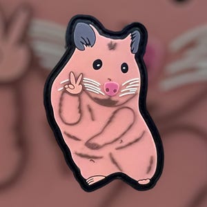 May include: A pink hamster with a black nose and white whiskers is giving a peace sign. The hamster is wearing a black outline and is standing on a pink background.