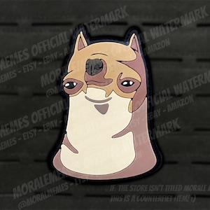 Puede incluir: Una criatura marrón y beige con aspecto de perro, con una expresión tonta. La criatura está representada en un estilo de dibujos animados con una cabeza grande y un cuerpo pequeño. La imagen es un parche con un borde negro.